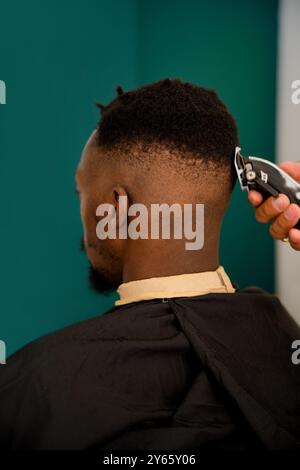 Ein professioneller Friseur schneidet sorgfältig die Haare eines schwarzen Mannes und zeigt eine trendige Frisur vor einem leuchtenden blaugrünen Hintergrund Stockfoto