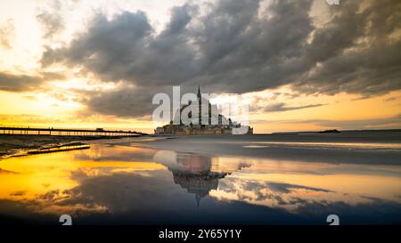 Ein ruhiger Sonnenaufgang wirft goldene Farbtöne über dem Mont Saint-Michel in der Normandie, dessen Reflexion sich perfekt im ruhigen Wasser unter einem Dramat spiegelt Stockfoto