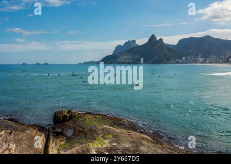 Rio de Janeiro, Brasilien. Blick auf den Strand Leblon und den Hügel Dois Irmãos von Arpoador an einem sonnigen Tag. Surfer im Meer warten auf die Welle. Stockfoto