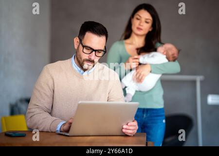 Der junge Vater arbeitet am Laptop, während seine Frau sich um ein Kleinkind kümmert. Arbeiten Sie von zu Hause aus Stockfoto