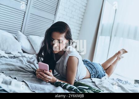 Meldungen werden geprüft. Schöne junge Frau, die ihr Smartphone hält und es mit einem Lächeln ansieht, während sie auf dem Bett liegt Stockfoto
