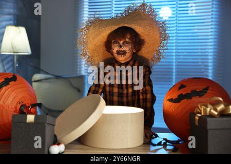 Süßer Junge, der wie Vogelscheuche gekleidet ist, mit festlichem Dekor und Geschenkboxen drinnen in der Nacht. Halloween-Feier Stockfoto