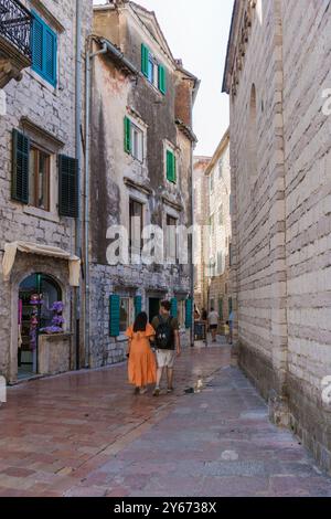 Wanderer erkunden die engen, kopfsteingepflasterten Straßen von Kotor, umgeben von alten Steinhäusern, die mit lebhaften Fensterläden geschmückt sind. Das Sonnenlicht taucht die historische Stadt in ein warmes Licht. Stockfoto