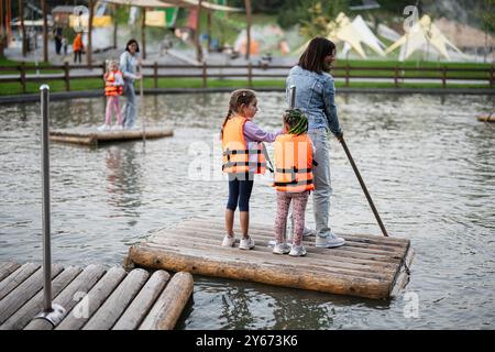 Zwei Kinder mit Schwimmwesten fahren unter Aufsicht eines Erwachsenen auf einem Teich. Die Szene spielt in einem Park, der eine sichere und abenteuerliche Umgebung bietet Stockfoto