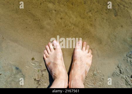 Blick von oben auf die Füße auf dem Sand, die auf eine Welle warten. Aus der Perspektive der ersten Person. Beine auf dem Hintergrund des Strandes Stockfoto