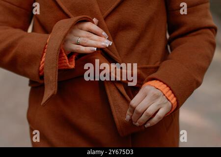 Bindegürtel für weibliche Hände auf einem warmen, braunen orangefarbenen Mantel, modische Details zur Herbstkleidung Stockfoto