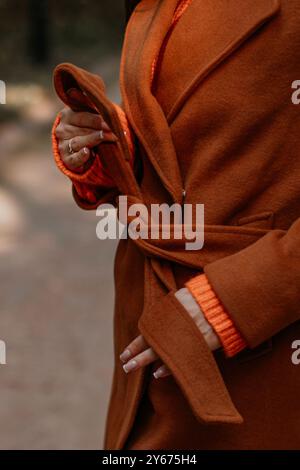 Bindegürtel für weibliche Hände auf einem warmen, braunen orangefarbenen Mantel, modische Details zur Herbstkleidung Stockfoto