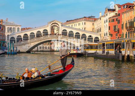 Ein Coupé, das eine Gondelfahrt macht, während ein Gondolierer seine Gondelfahrt entlang des Canal Grande in der Nähe der Rialtobrücke in Venedig, Italien, führt Stockfoto