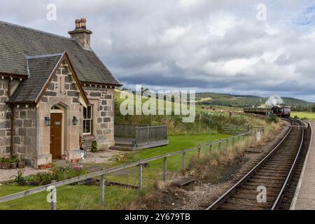 Die Strathspey Heritage Dampfeisenbahn verlässt den Bahnhof Broomhill, mit dem alten Haus des Bahnmeisters auf der linken Seite. Highlands, Schottland, Vereinigtes Königreich Stockfoto