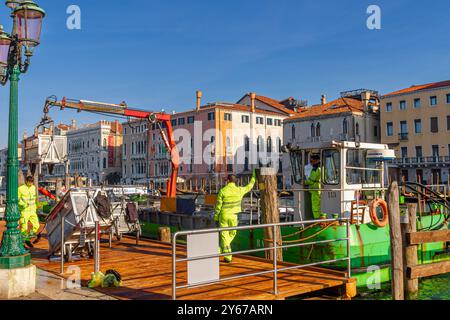 Männer auf einem Müllboot mit hydraulischem Hubarm sammeln und leeren Mülltonnen in der Nähe des Rialto-Markts entlang des Canale Grande in Venedig, Italien Stockfoto