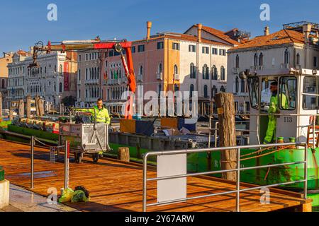 Männer auf einem Müllboot mit hydraulischem Hubarm sammeln und leeren Mülltonnen in der Nähe des Rialto-Markts entlang des Canale Grande in Venedig, Italien Stockfoto