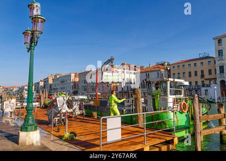 Männer auf einem Müllboot mit hydraulischem Hubarm sammeln und leeren Mülltonnen in der Nähe des Rialto-Markts entlang des Canale Grande in Venedig, Italien Stockfoto