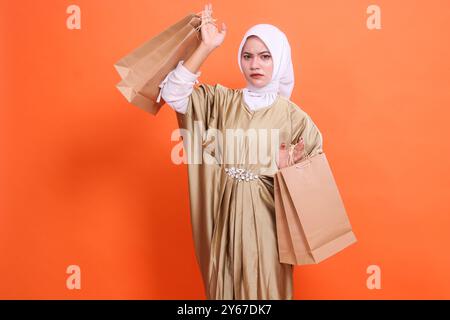 Wütende reife asiatische Frau mit brauner Papiertüte oben und unten, in der Lebensmittel im muslimischen Kaftan-Kleid mit Hijab stehen, isoliert auf orangefarbenem Backgrou Stockfoto