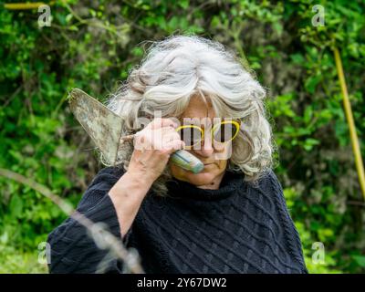 Eine Seniorin macht eine Pause von der Gartenarbeit, hält ihre Kelle und trägt eine gelbe Sonnenbrille, sieht zufrieden und glücklich aus, umgeben von Grün Stockfoto