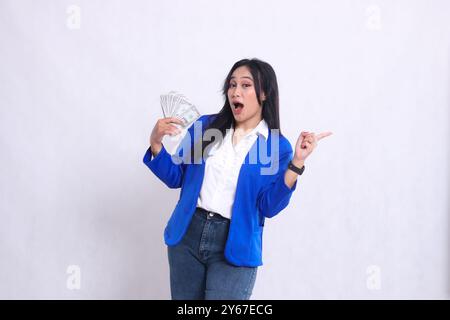 Schöne reife Bürofrau in blauem formellem Anzug, die überrascht vor der Kamera steht, Dollarscheine nach rechts hält und nach links zeigt, isoliertes weißes B Stockfoto