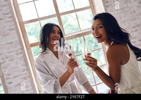 Braut mit bester Freundin. Zwei attraktive junge Frauen lächeln, während sie Champagner in der Garderobe trinken Stockfoto