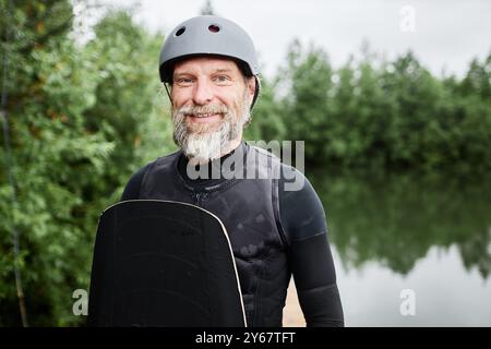 Porträt eines bärtigen älteren Mannes mit Wakeboardausrüstung und Helm, der draußen in die Kamera lächelt und am malerischen Waldsee steht Stockfoto