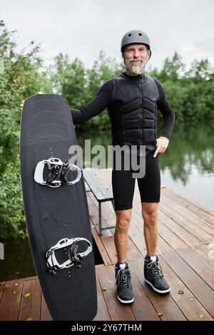 Vertikales Porträt eines bärtigen älteren Mannes, der Wakeboardausrüstung trägt und ein Brett hält, lächelnd in die Kamera, der draußen auf dem Pier am See steht Stockfoto