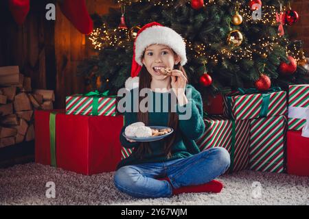 Großes Foto des süßen kleinen Mädchens sitzen Boden essen Snacks gekleidet grün weihnachten Outfit Tag Licht Haus Party Dekoration Innenraum Zimmer Stockfoto