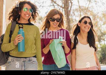 Drei Frauen lächeln und unterhalten sich, während sie draußen im Sonnenlicht laufen. Sie tragen legere Kleidung, Sonnenbrille und tragen umweltfreundliche Taschen und eine Wasserflasche Stockfoto
