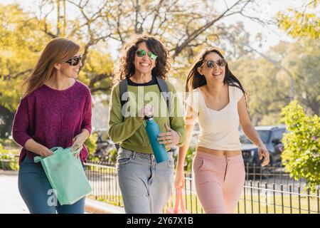 Eine Gruppe von drei Frauen, die in der Sonne spazieren und lachen, genießen einen entspannten Spaziergang im Park, mit Taschen und einer Flasche Wasser in der Hand. Das perfec Stockfoto