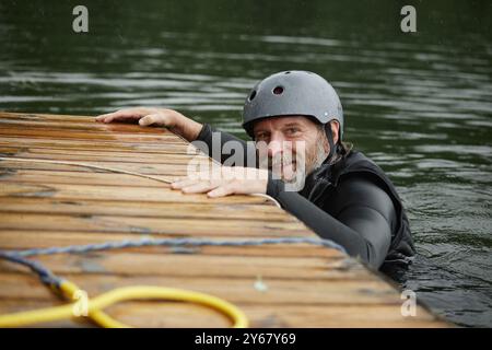 Porträt eines lächelnden bärtigen Mannes im Seewasser, der sich auf einen hölzernen Pier hält und in die Kamera lächelt, um Wassersport zu üben Stockfoto