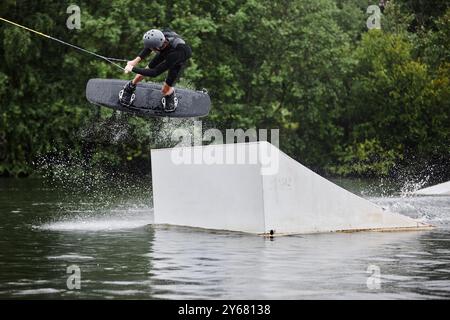 Weitwinkel-Action-Aufnahme von Sportlern, die Wakeboarden auf dem See genießen und hohe Sprünge auf der Rampe mit Wasserspritzern machen Stockfoto