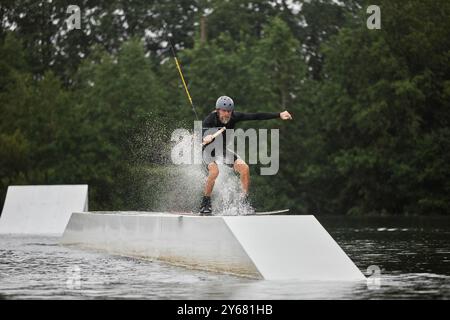 Weitwinkel-Action-Aufnahme eines sportlichen Seniorenmannes, der Wakeboarden genießt und Tricksprünge auf der Rampe macht, die sich auf die Kamera zubewegt, mit Spritzern Kopierraum Stockfoto