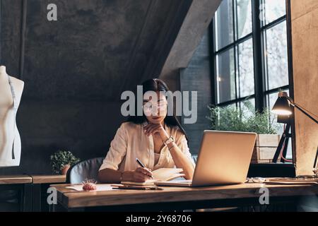 Und dann treffe ich mich mit... Konzentrierte junge Frau, die ihre Notizen im persönlichen Organisator überprüft, während sie in ihrer Werkstatt sitzt Stockfoto