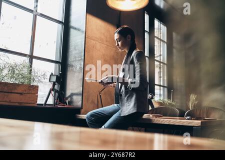 Immer online. Ernsthafte junge Frau in eleganter Freizeitkleidung mit digitalem Tablet, während sie sich auf den Schreibtisch lehnt Stockfoto