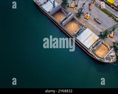 Luftaufnahme eines Frachtschiffes in einem Hafen, das während der Tageslichtstunden in einem stark frequentierten Hafen Körner hochlädt Stockfoto