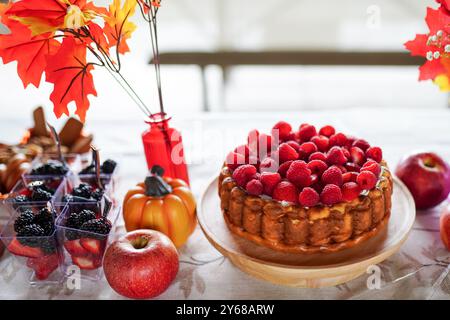 Ein festlicher Herbsttisch mit einem mit Himbeeren belegten Kuchen auf einem Holzständer, umgeben von Äpfeln, kleinen Kürbissen, Beerentassen und lebhaftem Herbstlaub Stockfoto