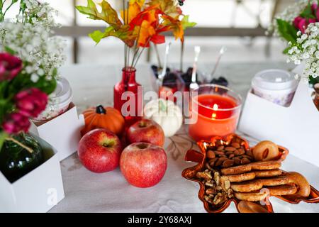 Ein gemütlicher Tisch im Herbststil mit Äpfeln, Kürbissen, Snacks und einer beleuchteten Kerze, umgeben von dekorativen Blumen und Herbstblättern Stockfoto