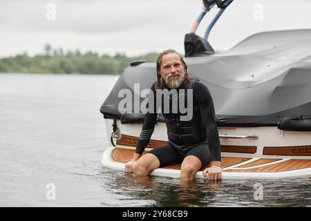 Porträt eines aktiven bärtigen Mannes, der einen Taucheranzug trägt und in die Kamera lächelt, während er auf der Rückseite des Bootes sitzt, nachdem er im Kopierraum des Sees geschwommen ist Stockfoto