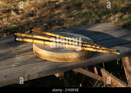 Drei Kaminflöten ruhen auf einer traditionellen Rahmentrommel auf einem rustikalen Holztisch, der in einem friedlichen Olivenhain unter natürlichem Licht steht Stockfoto