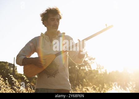 Ein Mann im Alter von 40 Jahren, der ein weißes Hemd trägt und das traditionelle baglama-Instrument in einem ruhigen Olivenfeld spielt, beleuchtet vom sanften Schein der setti Stockfoto