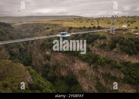 Ein atemberaubender Blick aus der Luft auf eine Diamond Bridge aus Glas, die über einem tiefen Canyon hängt, umgeben von zerklüfteten Klippen und üppigem Grün unter einem bewölkten Himmel. Stockfoto