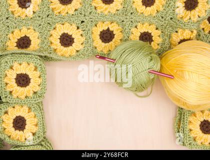 Nahaufnahme von Häkeln von fertig gehäkelten Oma-Quadraten, verbundene Quadrate mit Kugeln aus gelbem und grünem Garn zur Seite auf hellem Holztisch. Croche Stockfoto