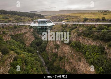 Ein atemberaubender Blick aus der Luft auf eine Diamond Bridge aus Glas, die über einem tiefen Canyon hängt, umgeben von zerklüfteten Klippen und üppigem Grün unter einem bewölkten Himmel. Stockfoto