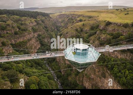 Ein atemberaubender Blick aus der Luft auf eine Diamond Bridge aus Glas, die über einem tiefen Canyon hängt, umgeben von zerklüfteten Klippen und üppigem Grün unter einem bewölkten Himmel. Stockfoto