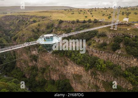 Ein atemberaubender Blick aus der Luft auf eine Diamond Bridge aus Glas, die über einem tiefen Canyon hängt, umgeben von zerklüfteten Klippen und üppigem Grün unter einem bewölkten Himmel. Stockfoto