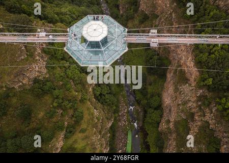 Ein atemberaubender Blick aus der Luft auf eine Diamond Bridge aus Glas, die über einem tiefen Canyon hängt, umgeben von zerklüfteten Klippen und üppigem Grün unter einem bewölkten Himmel. Stockfoto