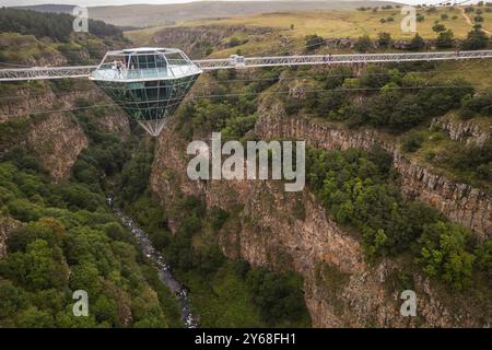 Ein atemberaubender Blick aus der Luft auf eine Diamond Bridge aus Glas, die über einem tiefen Canyon hängt, umgeben von zerklüfteten Klippen und üppigem Grün unter einem bewölkten Himmel. Stockfoto