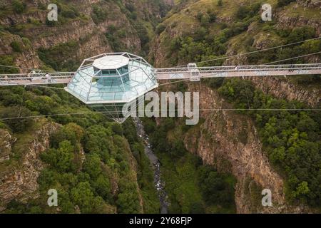 Ein atemberaubender Blick aus der Luft auf eine Diamond Bridge aus Glas, die über einem tiefen Canyon hängt, umgeben von zerklüfteten Klippen und üppigem Grün unter einem bewölkten Himmel. Stockfoto