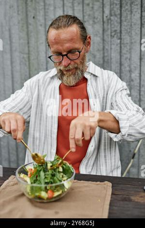 Vertikales Porträt des bärtigen Reifen Mannes, der legere Outfit trägt, Salat beim Genießen eines gesunden Mittagessens im Café im Freien Stockfoto