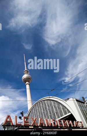 Das Bahnhofsschild Alexanderplatz steht mit dem Fernsehturm, der sich vor einem hellblauen Himmel in Berlin erhebt. Stockfoto