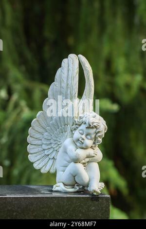 Kleine weiße Engelsstatue auf einem Grabstein auf einem Friedhof. Fichtenzweige im Hintergrund. Das Bild hat einen leeren Platz für Text. Stockfoto
