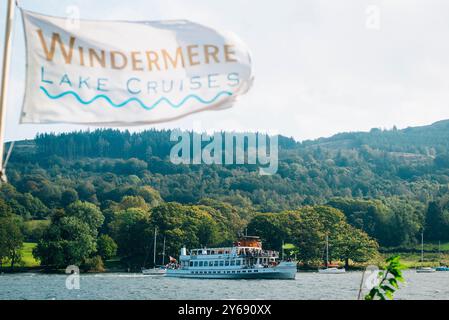 Eine Fähre fährt auf Windermere Lake Cruises im Lake District mit einer Flagge, die im Wind weht, umgeben von Hügeln, Bäumen und friedlichem Wasser. Stockfoto