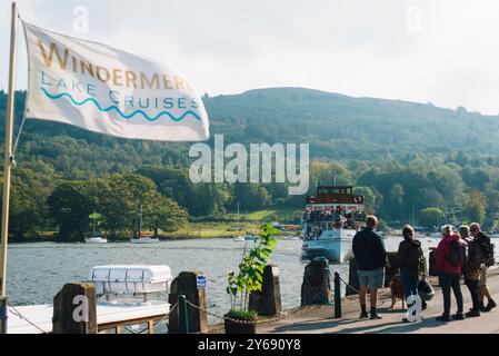 Touristen warten am Dock auf ein Boot auf den Windermere Lake Cruises im Lake District an einem windigen Tag, umgeben von Hügeln, Bäumen und friedlichem Wasser. Stockfoto