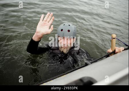 Filmporträt eines fröhlichen bärtigen Mannes, der in den See fällt und vor der Kamera winkt und Wassersport mit dem Boot im Kopierraum genießt Stockfoto